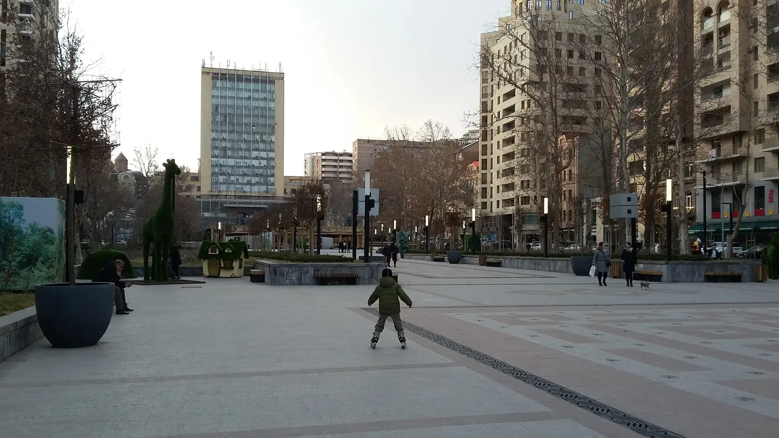 Un large boulevard piéton pavé en hiver, avec des topiaires, des lampadaires et un enfant en rollers, menant vers la tour de Haypost à l’arrière-plan