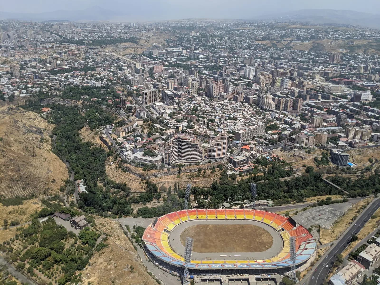 Vue aérienne d’Erevan en été : au premier plan l’ovale du stade Hrazdan et la gorge boisée, sur le rebord des tours et le quartier bas de Kond, et derrière le centre densément bâti