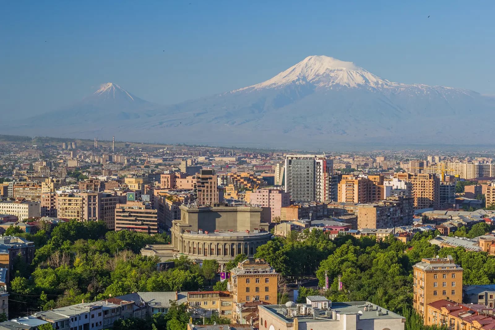 Vue en hauteur vers le sud sur le centre d’Erevan, avec l’Opéra circulaire au milieu des arbres, des immeubles de hauteur moyenne et une haute tour grise, et derrière le Grand et le Petit Ararat enneigés