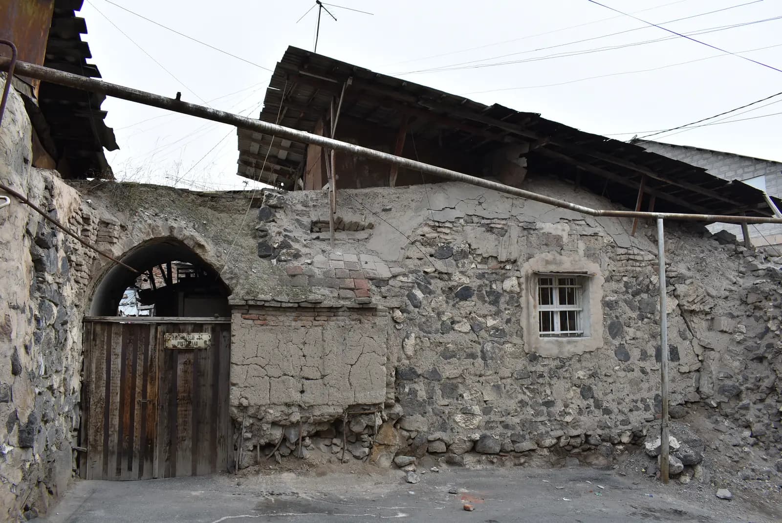 Un vieux mur de moellons de basalte, de briques et d’enduit de terre avec un portail en bois en arc brisé portant une plaque de rue bilingue, des toits de tôle affaissés au-dessus et un tuyau de gaz rouillé en travers de l’image