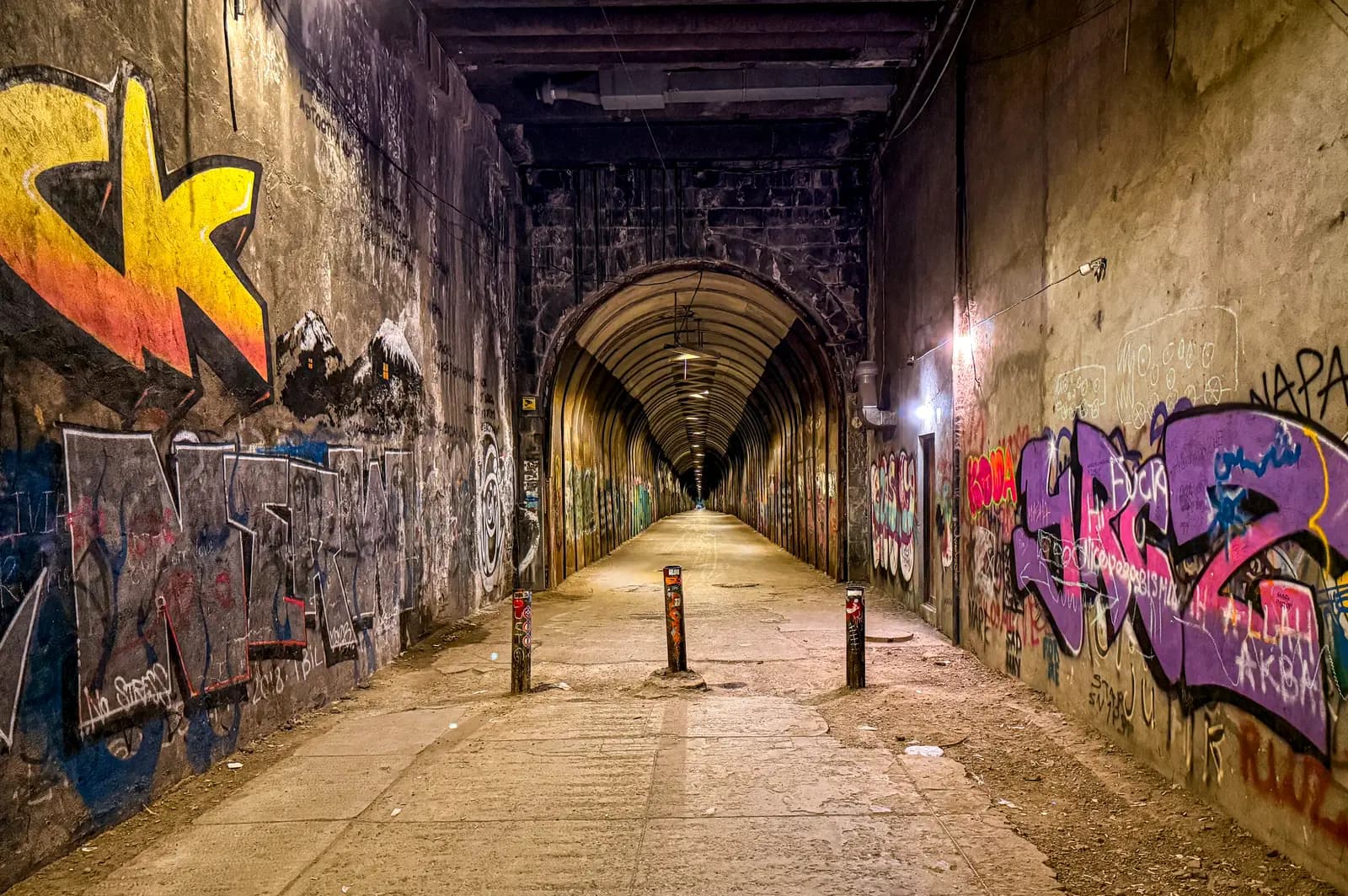 Un long tunnel piéton aux murs couverts de graffitis, des bornes au premier plan et un passage voûté éclairé qui s’étire au loin