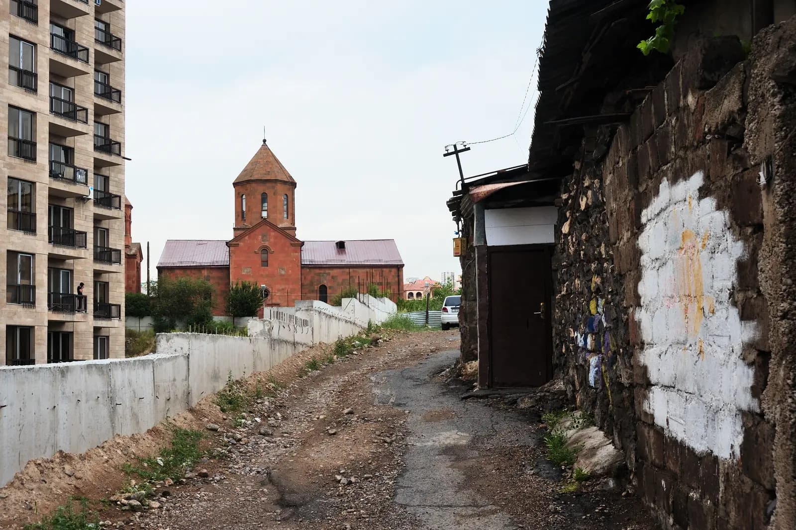 Un chemin de terre défoncé entre un mur de moellons de basalte délabré avec un abri à droite et un mur de béton à gauche sous un immeuble beige neuf, menant à l’église en tuf rouge Surb Hovhannes