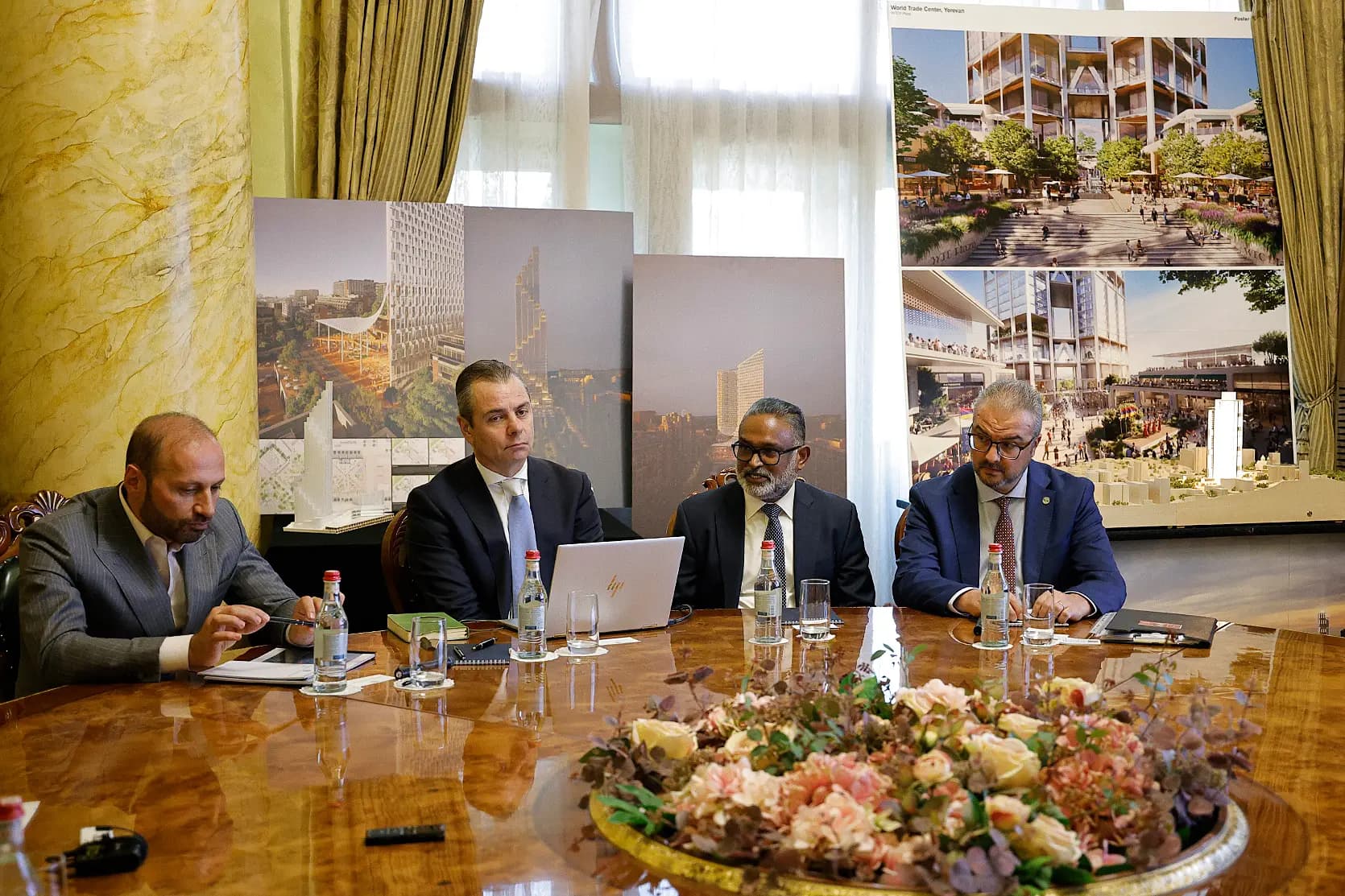Quatre hommes en costume autour d’une table ronde dans une salle à colonne de marbre et rideaux dorés ; derrière eux, de grands panneaux imprimés montrant une haute tour blanche et une place paysagée aux larges marches – l’un intitulé « World Trade Center, Yerevan » et marqué « Foster » – ainsi qu’une petite maquette blanche de tour sur un support