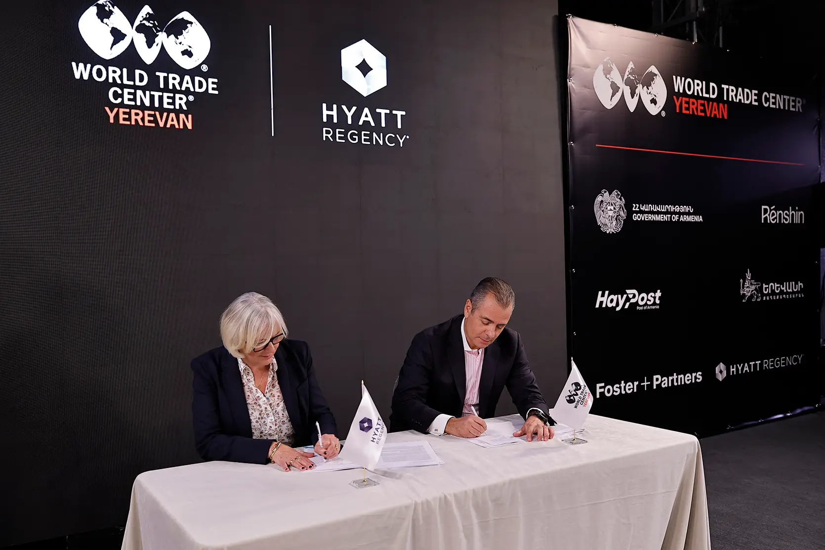 Deux personnes signent des documents à une table nappée de blanc, avec des fanions de table Hyatt Regency et WTC Yerevan, devant un écran affichant les logos de World Trade Center Yerevan et de Hyatt Regency