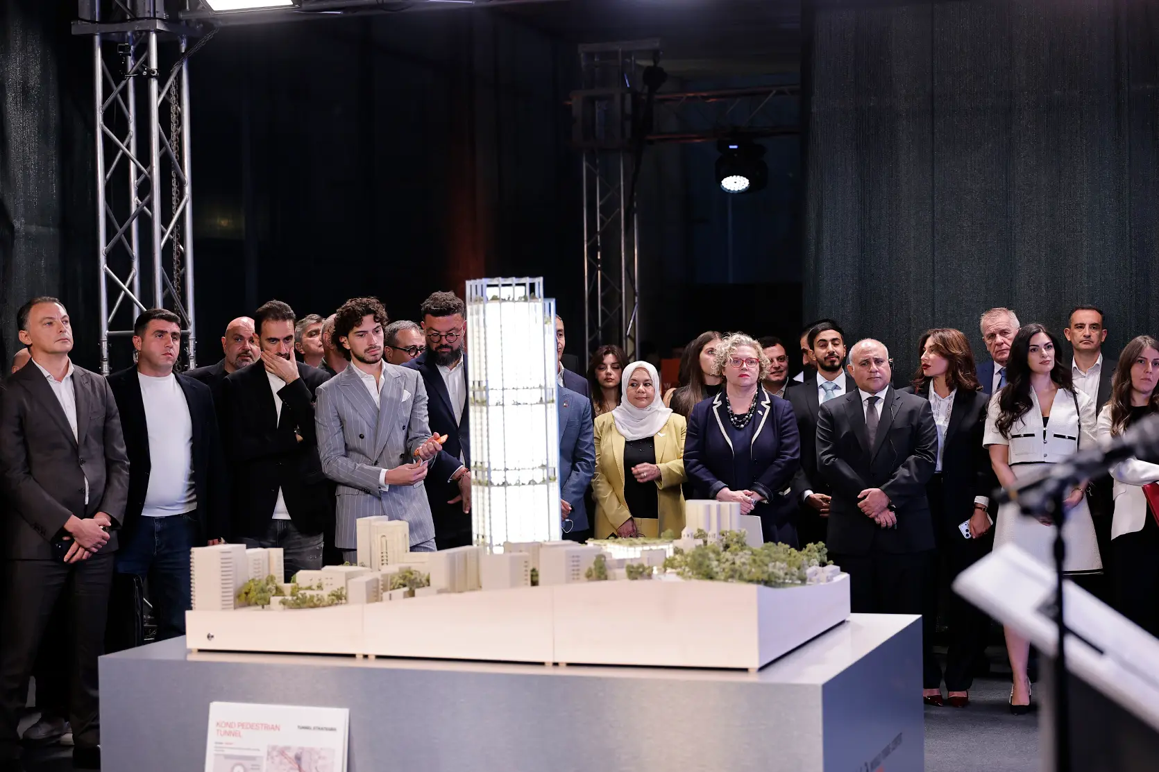 Vue de face d’une longue maquette blanche sur un socle gris : une maquette des tours éclairée de l’intérieur s’élève parmi des volumes blancs et des espaces plantés, un panneau intitulé « Kond Pedestrian Tunnel » est appuyé contre le socle et une rangée d’invités se tient derrière