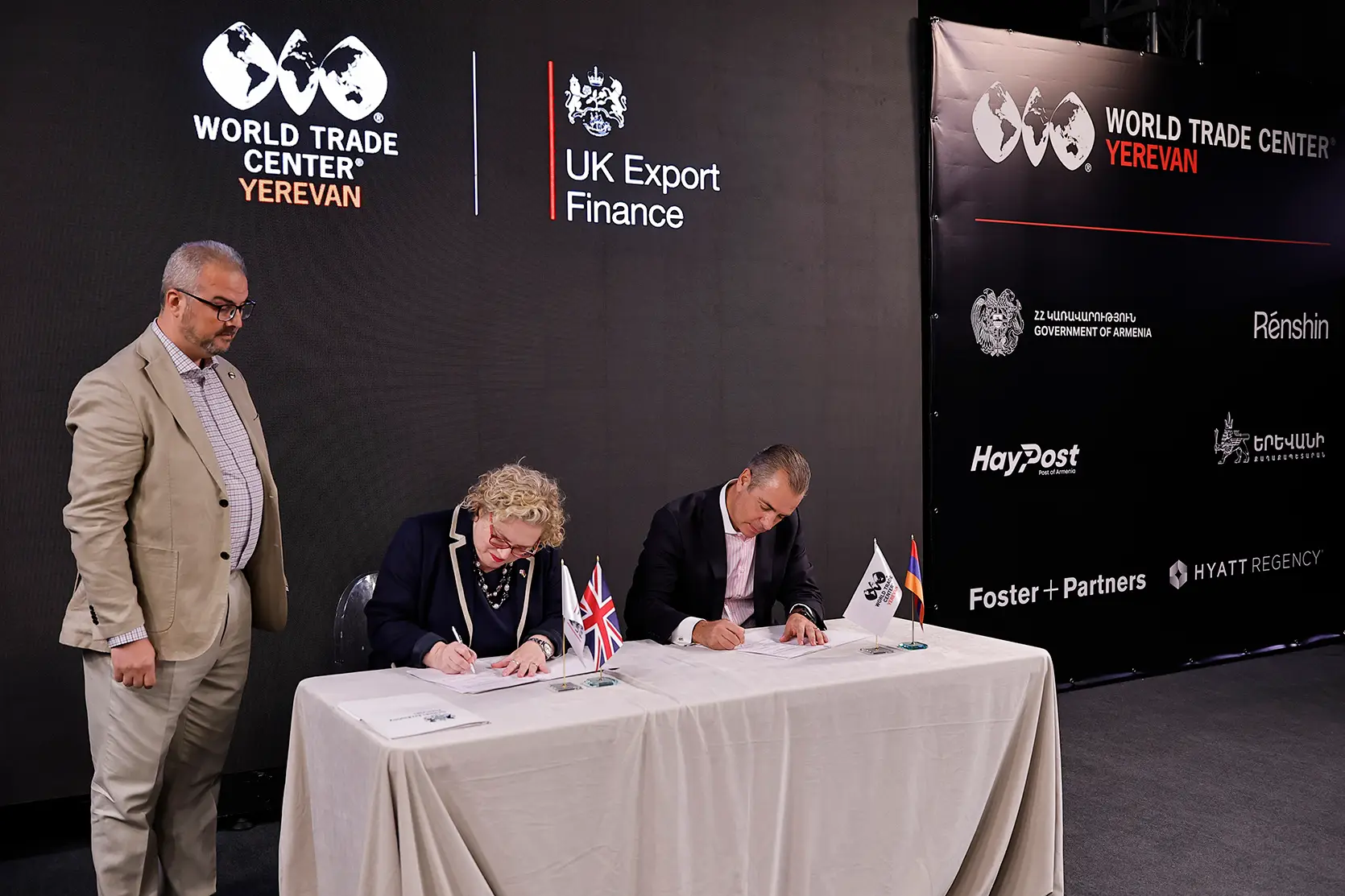 Deux personnes signent des documents à une table portant de petits drapeaux britannique et arménien et des fanions WTC Yerevan, sous le regard d’un homme en costume beige, devant un écran affichant les logos de World Trade Center Yerevan et de UK Export Finance