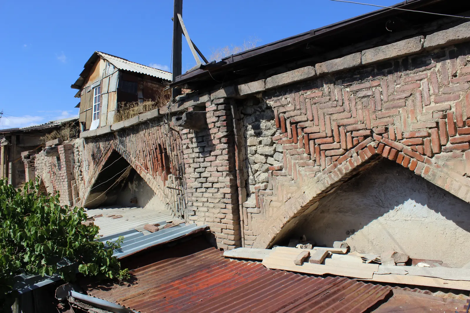 Vieux mur de brique à grands arcs brisés et appareil en arête de poisson, toits de tôle ondulée rouillée des maisons adossées à sa base et pièce en bois sur le dessus, sous un ciel bleu