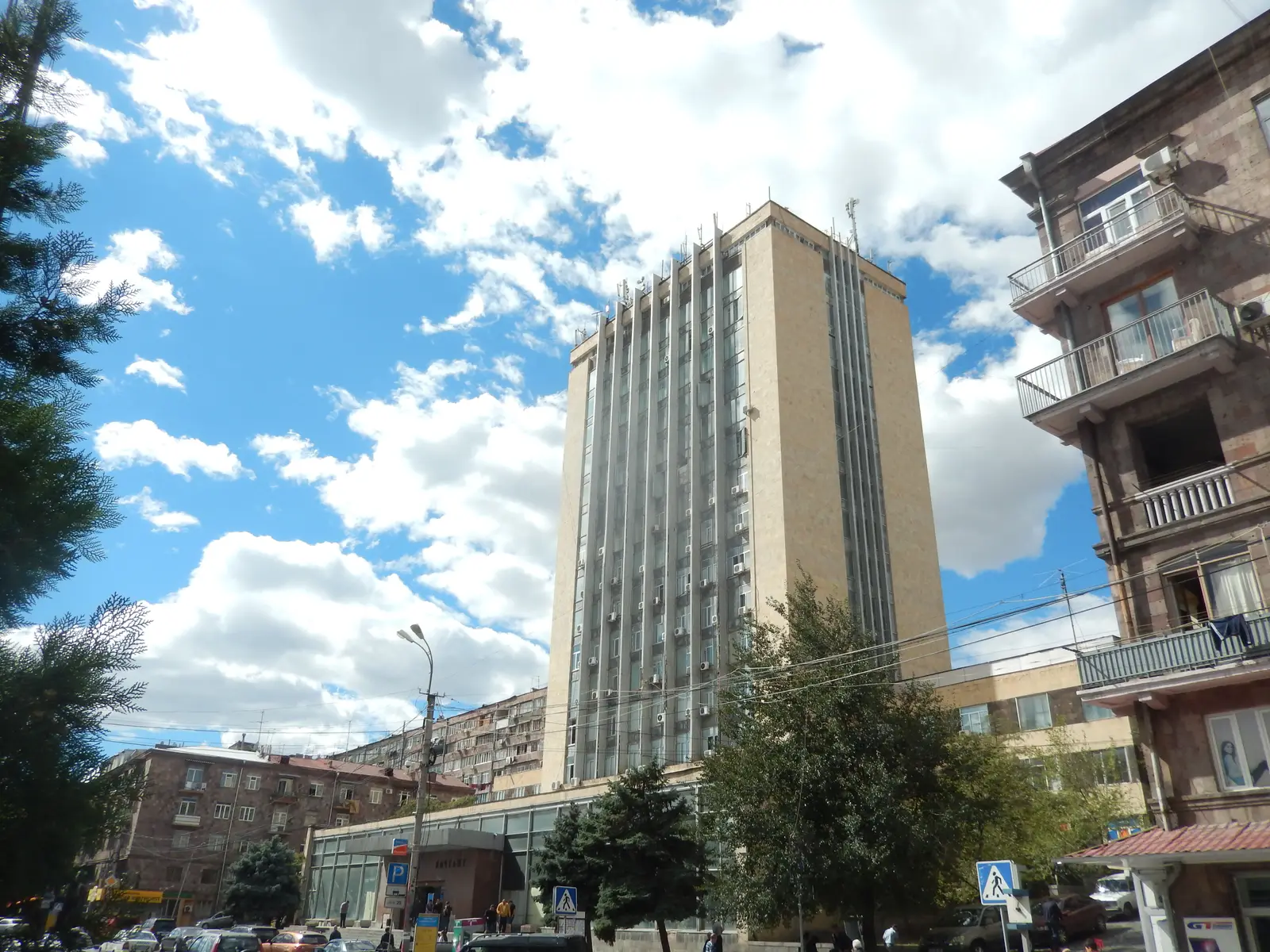 L’ancienne tour du siège de Haypost au 22 rue Saryan sous un ciel bleu nuageux : bâtiment en tuf clair à ailettes verticales et climatiseurs, avec au premier plan des arbres et un immeuble d’habitation en tuf rose