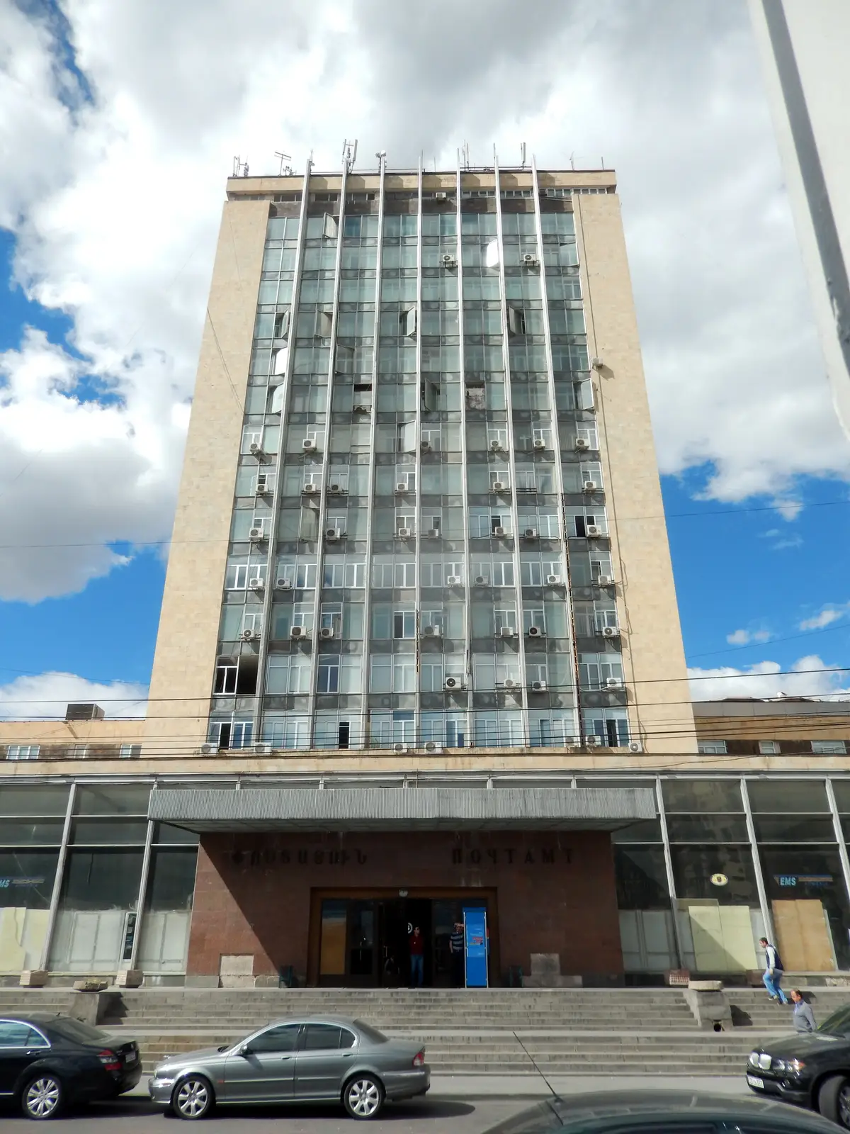 Façade du bâtiment de Haypost au 22 rue Saryan vue d’en bas : façade vitrée à ailettes verticales au-dessus d’un long rez-de-chaussée vitré, entrée en granit rouge portant l’inscription « ՓՈՍՏԱՏՈՒՆ / ПОЧТАМТ », marches et voitures garées