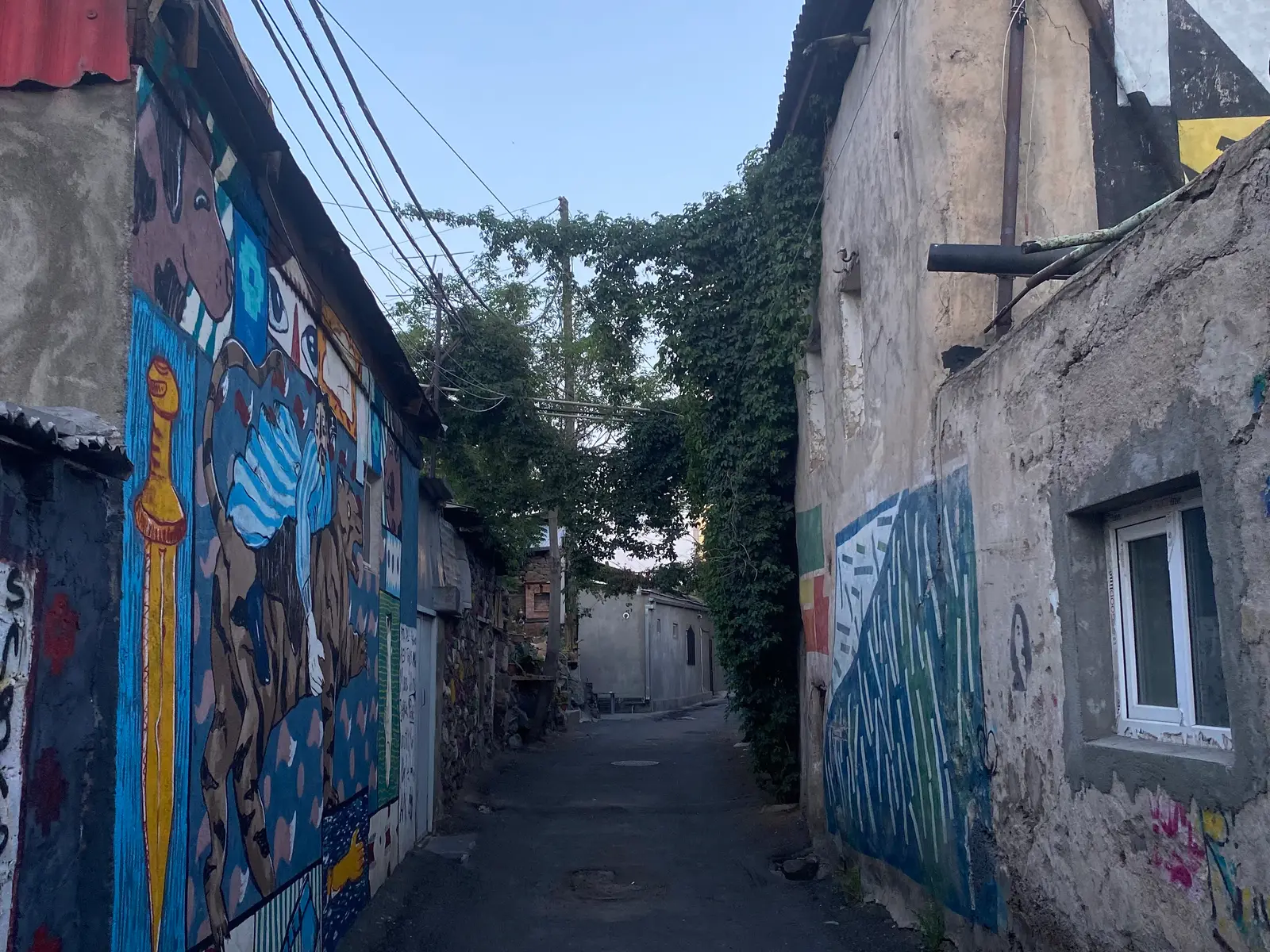 Une ruelle étroite de Kond entre de vieilles maisons d’un ou deux niveaux couvertes de peintures murales colorées de style populaire, sous une arche de lierre et des fils enchevêtrés