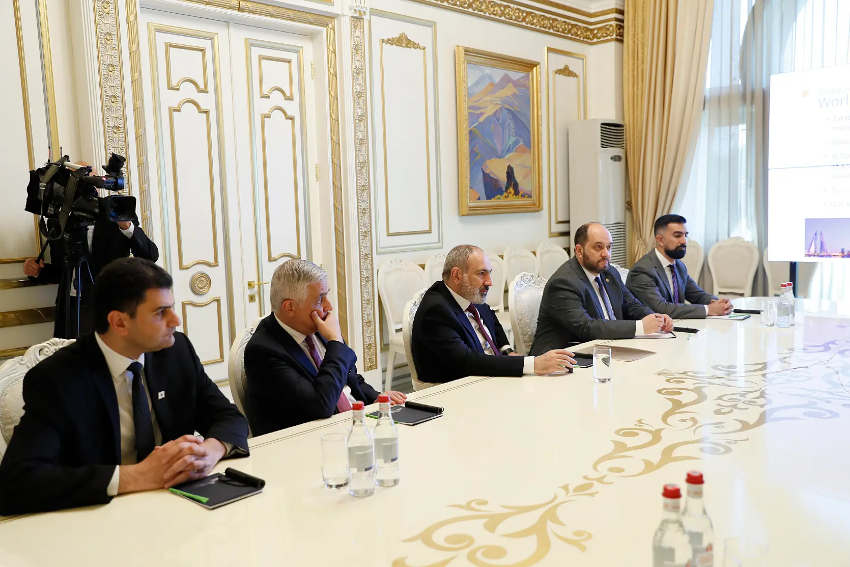 Le Premier ministre Nikol Pashinyan et quatre autres hommes en costume, assis à une table claire ornée de motifs dorés dans une salle blanc et or, regardent une présentation projetée sur un écran au bord droit de l’image, tandis qu’un caméraman filme