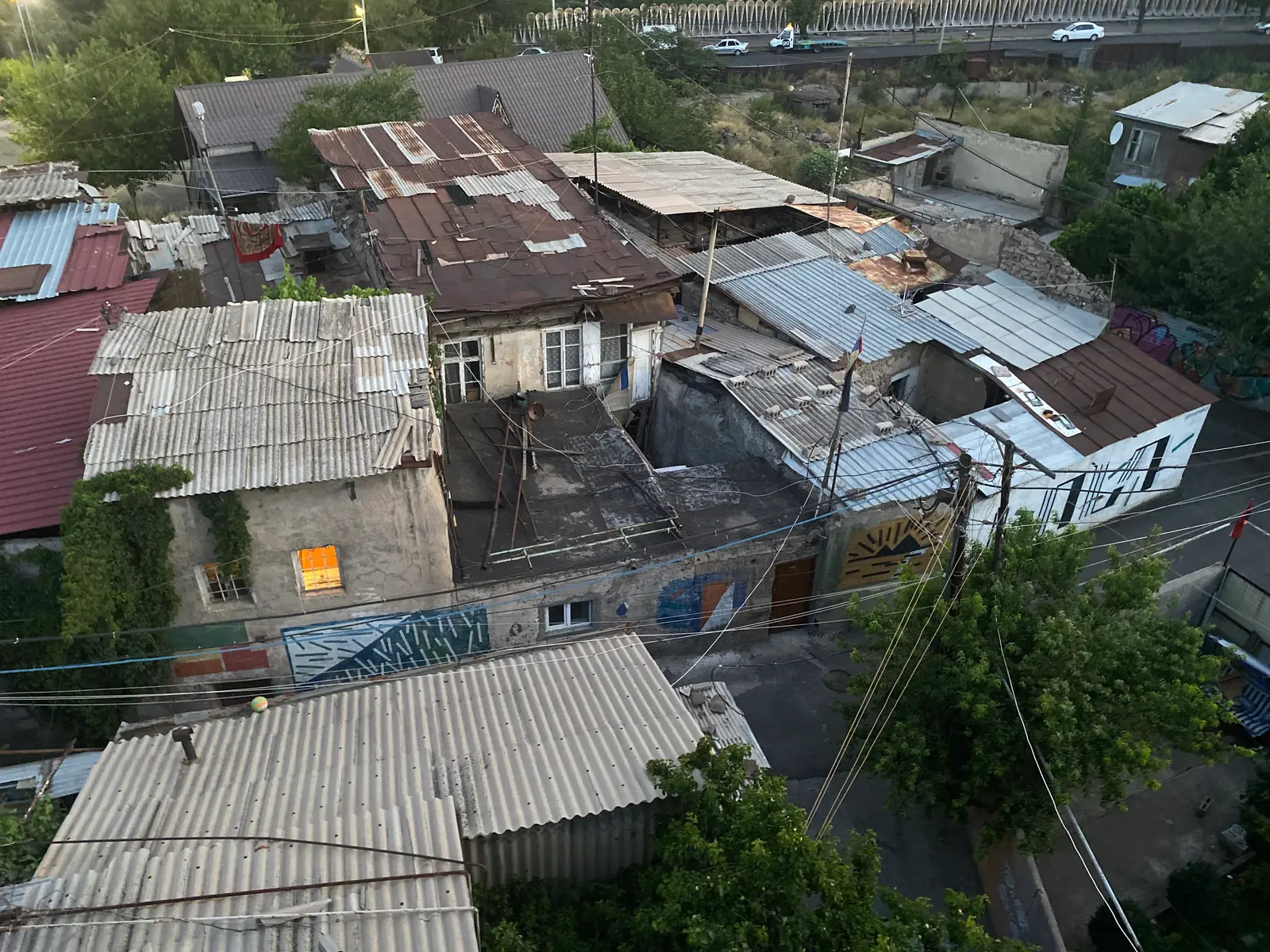Vue plongeante sur de vieilles maisons de plain-pied à Kond : toits de tôle ondulée rapiécés, peintures murales géométriques, fils aériens enchevêtrés et arbres, avec en haut une route bordée d’une balustrade